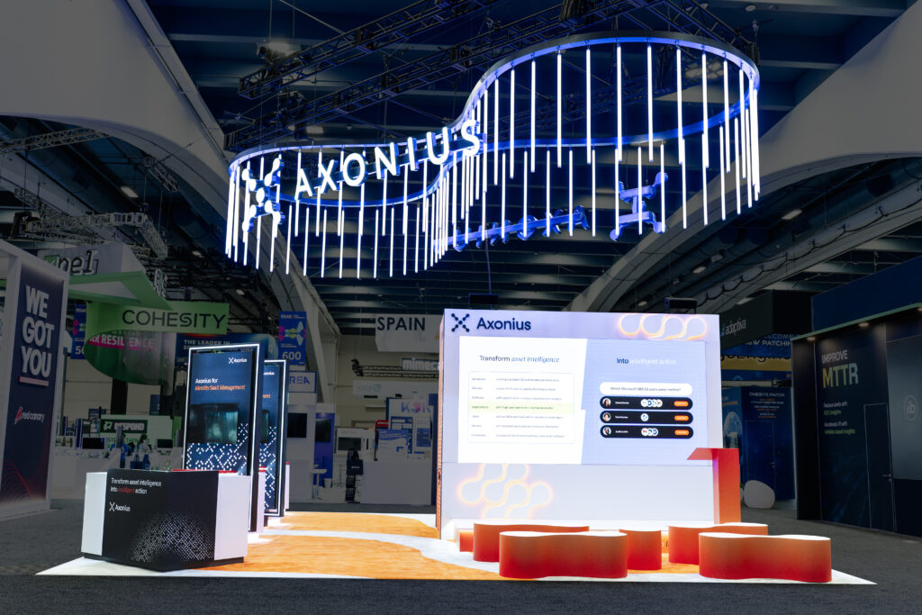 Trade show booth featuring a large overhead LED light installation, backlit signage, digital display panels, and a bright orange carpet in a well-lit convention hall.