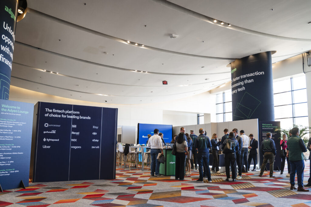 Wide shot of lounge area at a trade show with branded signage, networking attendees, and multiple booth activations under a modern, curved ceiling