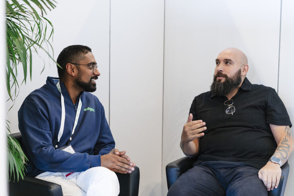 Two professionals in conversation seated in a casual expo lounge setting, surrounded by greenery and soft lighting at a corporate event