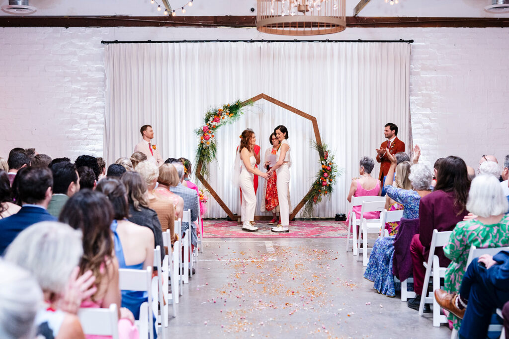 Couple celebrating their wedding ceremony with family and friends in a modern indoor venue, illustrating the community-focused experience of a traditional wedding.