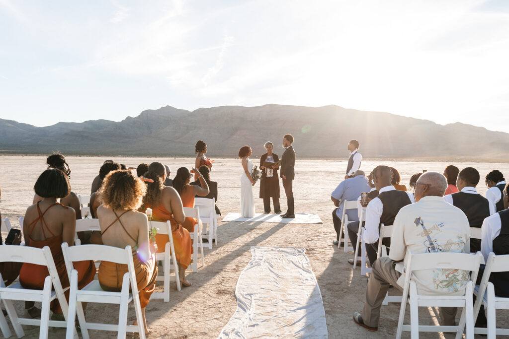 Las Vegas intimate wedding photographer captures couple exchanging vows at golden hour on the Jean Dry Lake Bed, bride in a fitted white gown, groom in a black suit, surrounded by seated guests in warm earth tones with the Nevada mountains glowing in the backlit sunset