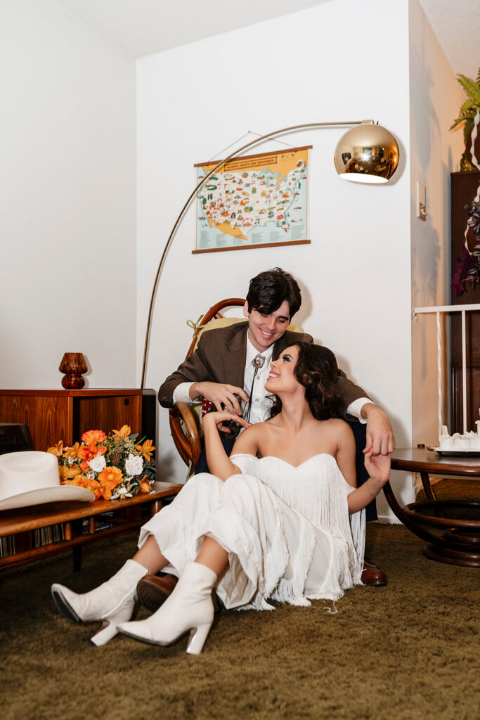 Las Vegas elopement photographer captures bride in a white fringe dress and white cowboy boots seated on the floor leaning against her groom in a brown suit at That 70s House, surrounded by retro mid-century furniture, a vintage arc lamp, and an orange floral bouquet