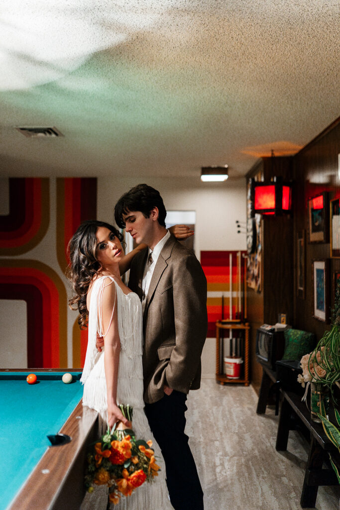 Las Vegas elopement photographer captures bride in a white fringe dress holding an orange bouquet leaning into her groom in a brown blazer beside a turquoise pool table, with iconic retro orange and red striped wallpaper in the background at That 70s House