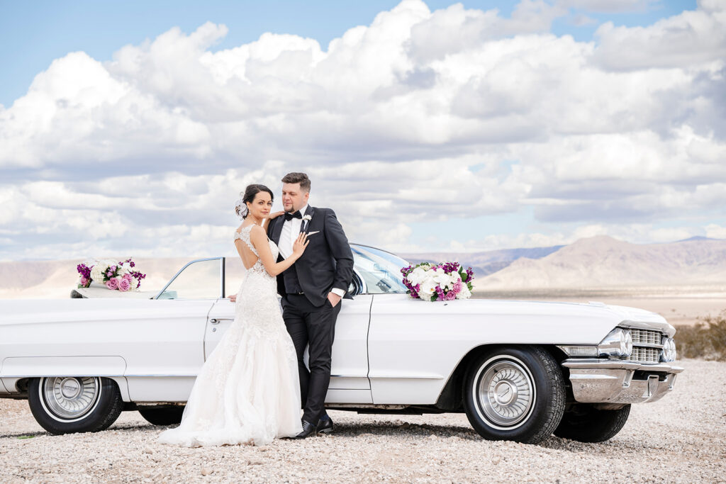 Newlyweds posing beside a classic white vintage car during a Las Vegas desert elopement portrait session at a dry lake bed with mountains in the background.