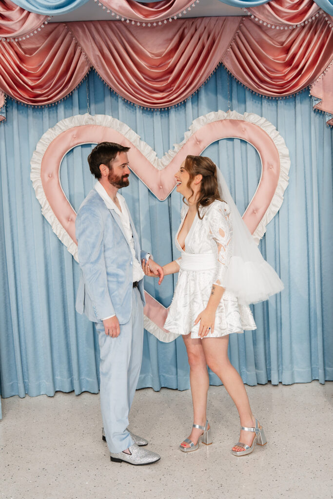 Las Vegas intimate wedding photographer captures couple laughing and holding hands in front of the iconic pink and blue heart backdrop at Sure Thing Chapel, bride in a short lace dress and veil, groom in a light blue suit with silver glitter shoes