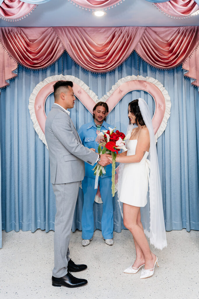 Couple exchanging vows at Las Vegas chapel ceremony with Elvis officiant and heart backdrop — elopement photography by True Colors Creative