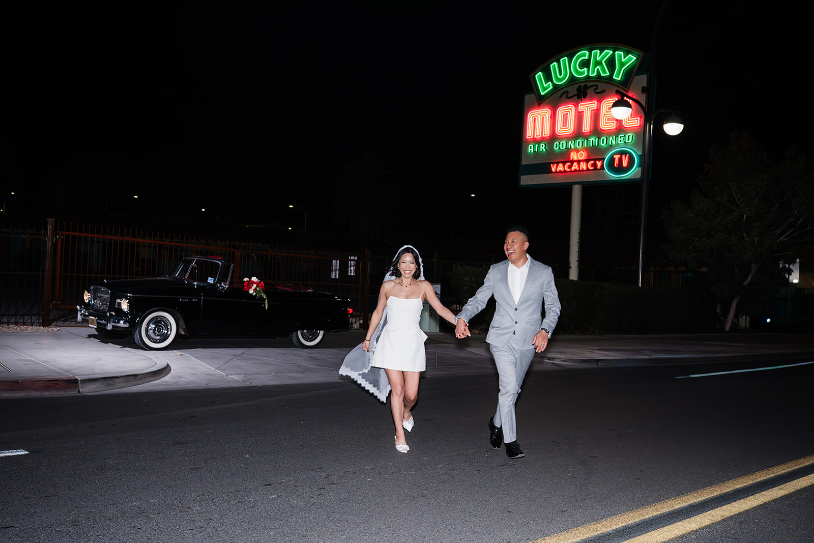 Bride and groom running hand in hand across a Las Vegas street at night beneath the Lucky Motel neon sign during their vintage-inspired elopement.