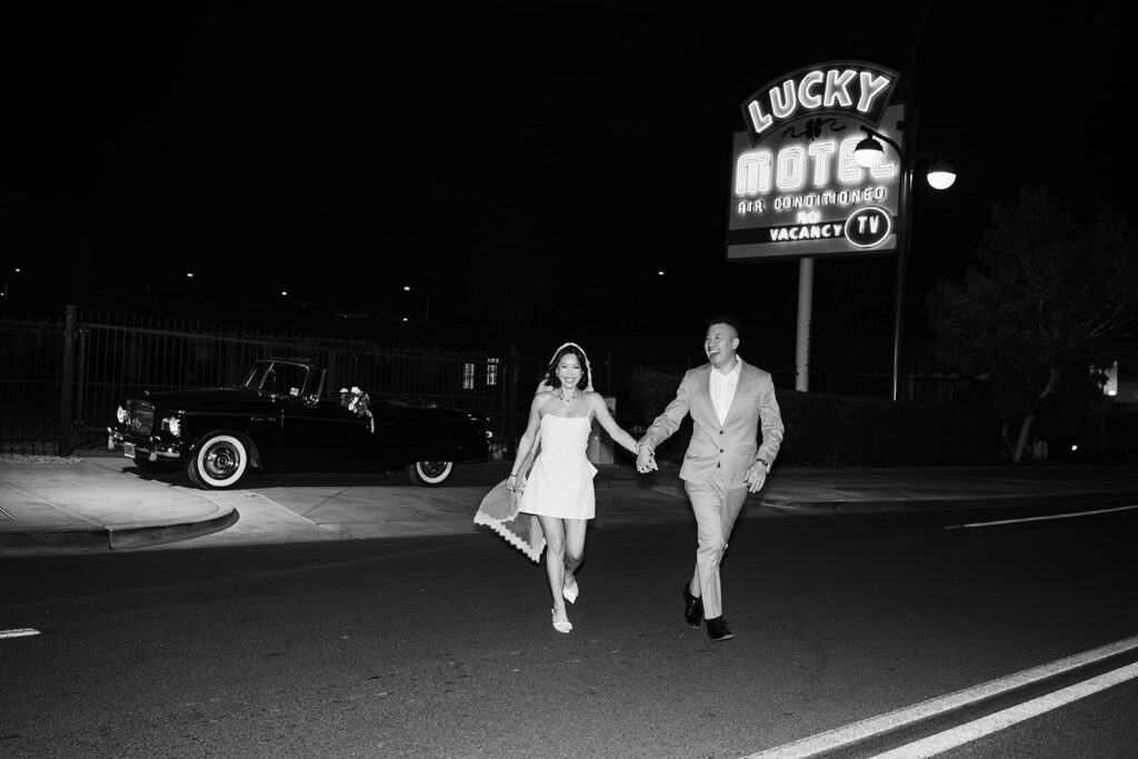 Fremont Street elopement photographer captures couple joyfully running past the Lucky Motel neon sign at night, bride in a modern short wedding dress, vintage classic car parked behind them — Las Vegas intimate wedding photography