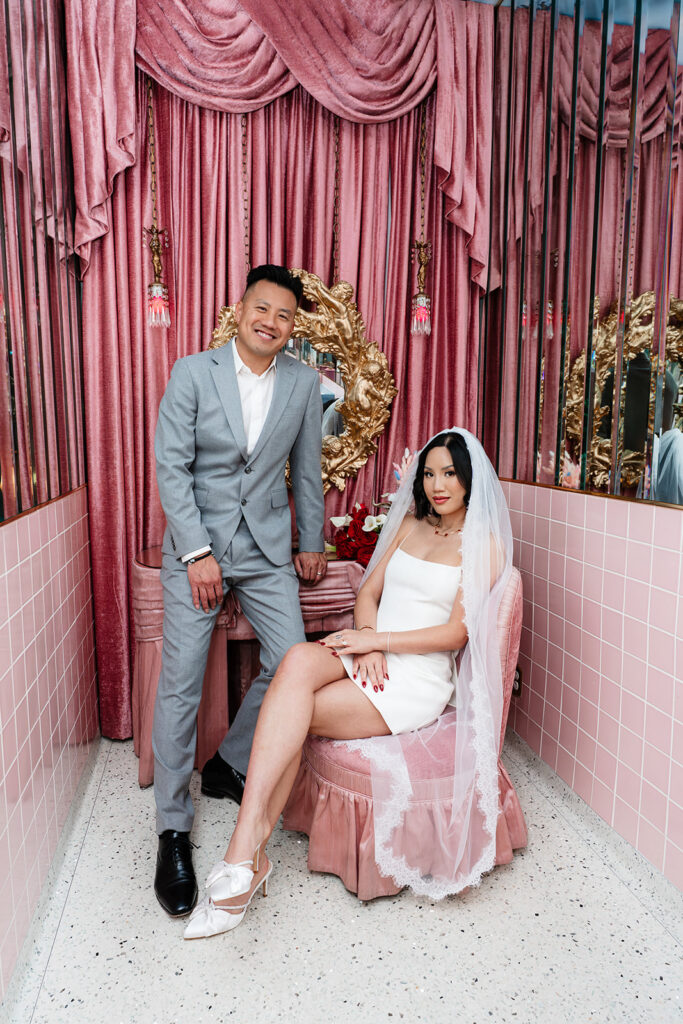 Las Vegas elopement photographer captures bride in a short white dress and veil seated on a pink velvet ottoman beside her groom in a grey suit, inside the iconic pink tiled room at Sure Thing Chapel
