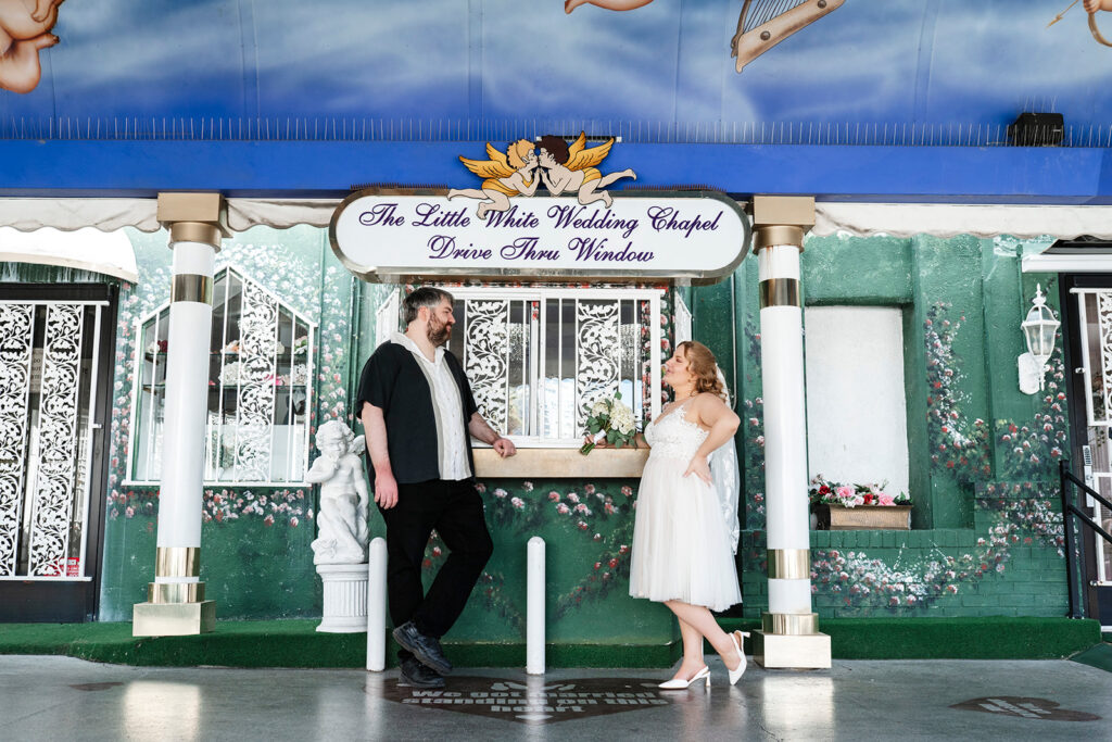 Las Vegas elopement photographer captures couple sharing a laugh outside the iconic Little White Wedding Chapel Drive Thru Window, bride in a midi-length white dress and veil holding a bouquet, groom in a casual black shirt