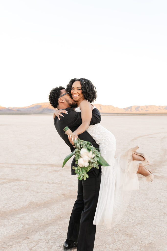 Groom lifting bride on the Las Vegas dry lake bed at sunset, Red Rock mountains in the background — elopement photography by True Colors Creative
