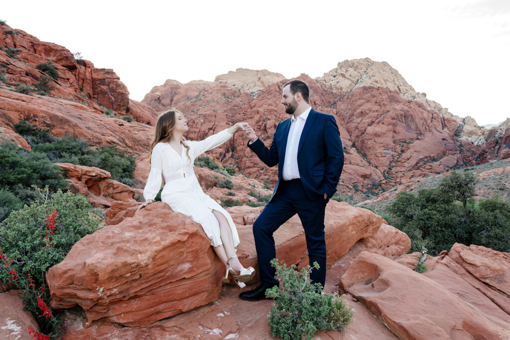 Red Rock Canyon elopement photographer captures bride in a long white boho dress seated on a red sandstone rock holding hands with her groom in a navy suit, surrounded by dramatic desert rock formations and desert brush