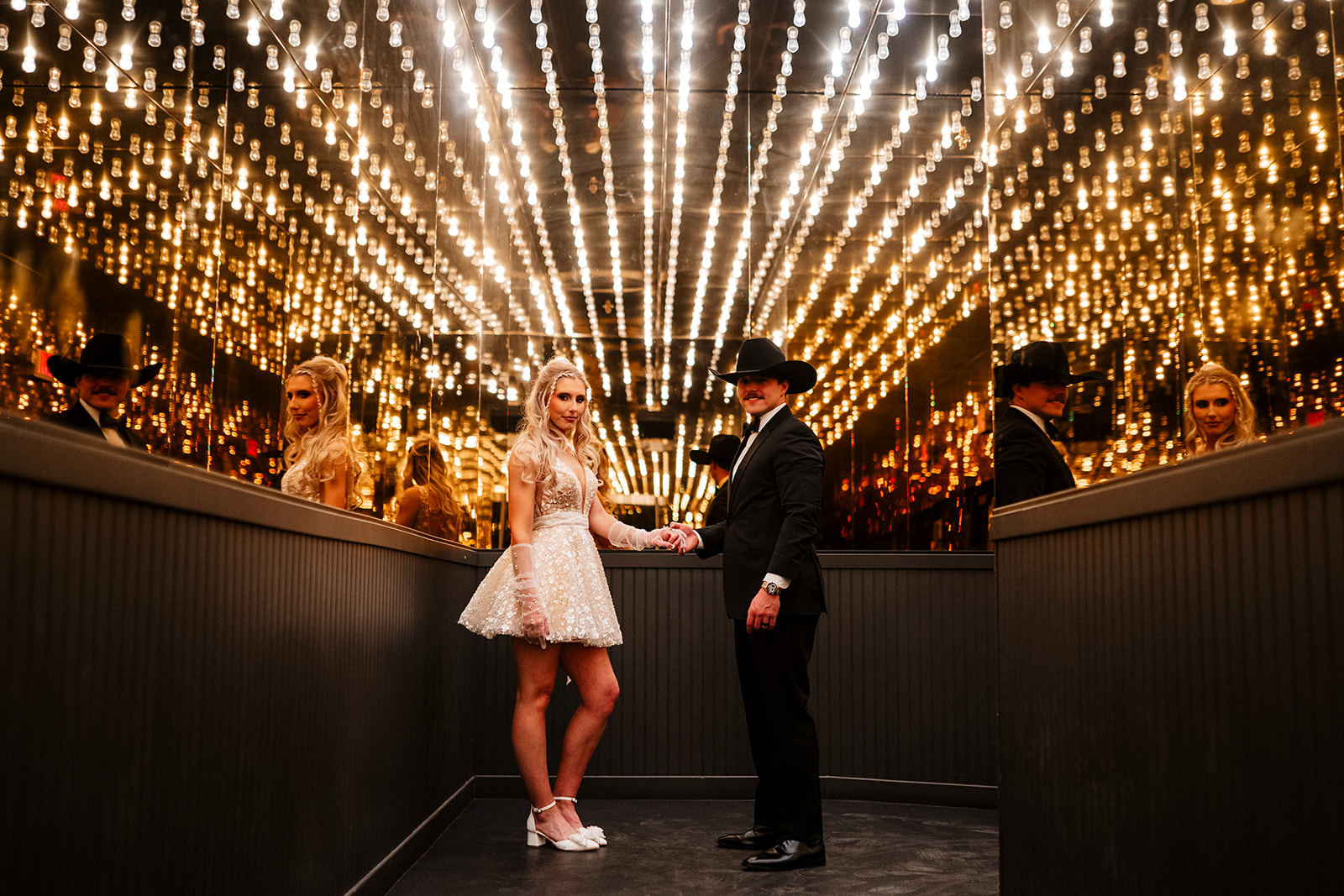 Las Vegas elopement photographer captures bride in a short sparkly dress and groom in a black tux and cowboy hat holding hands in a mirrored room with hundreds of glowing Edison bulbs — True Colors Creative