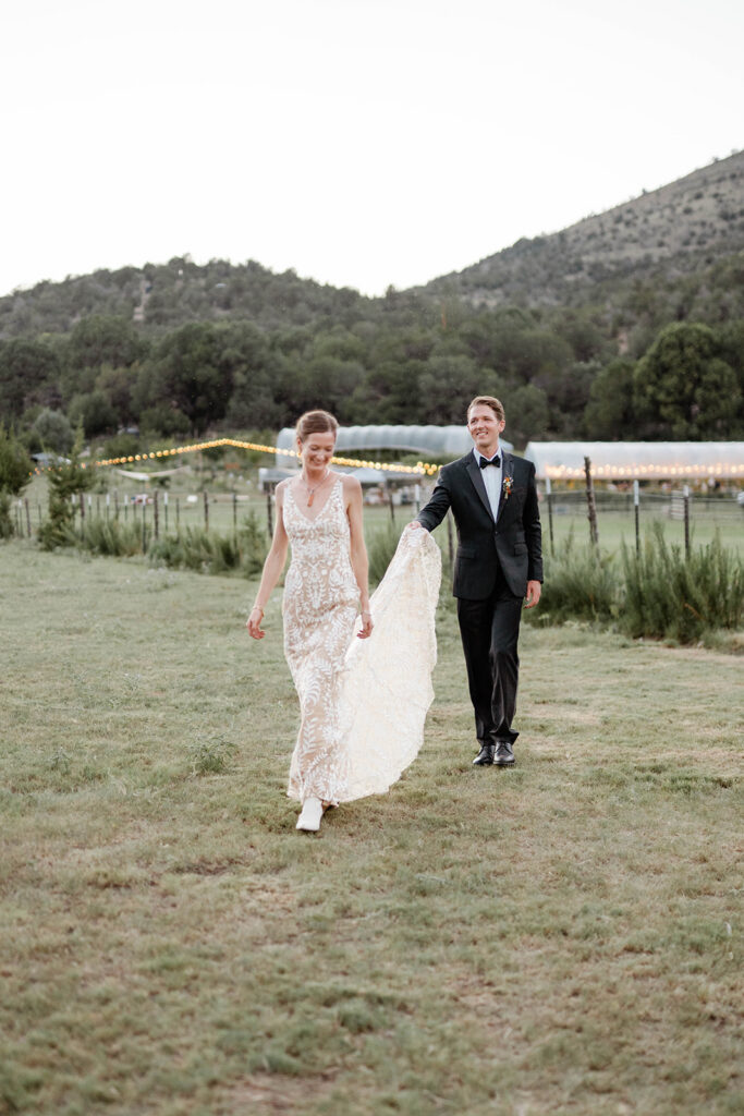 Destination wedding photographer captures bride and groom walking through open field at dusk with string lights and mountain backdrop at outdoor ranch wedding in the Southwest
