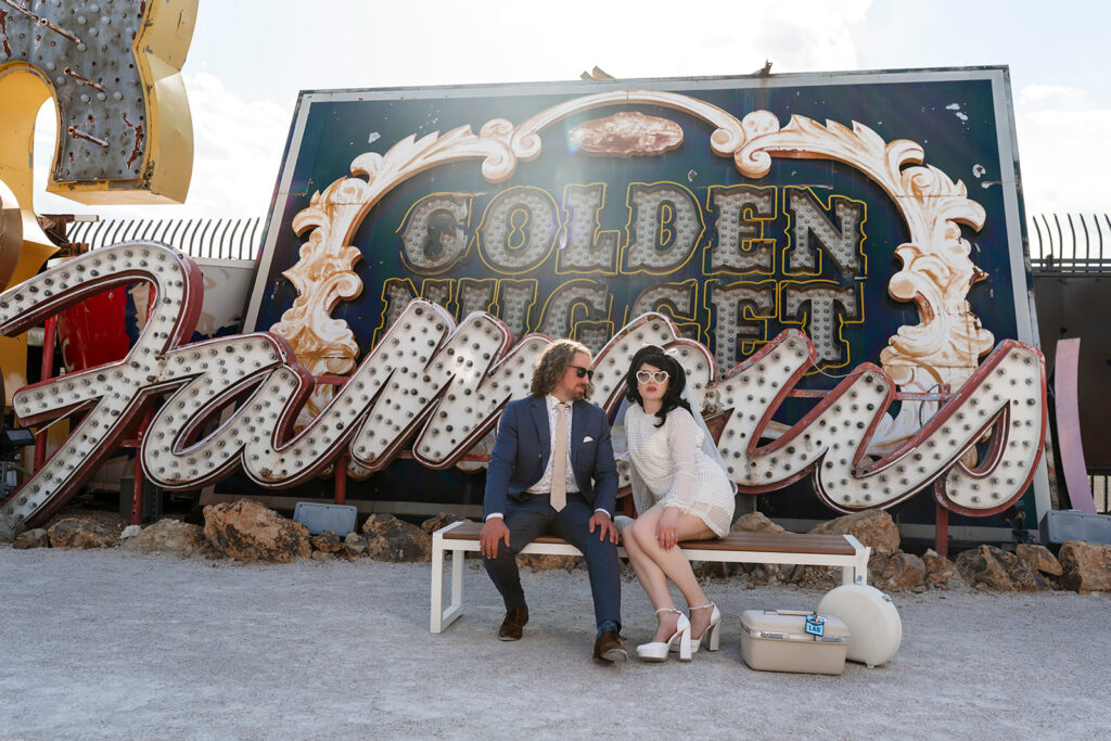 Neon Museum elopement photographer captures couple seated on a bench in the Neon Boneyard, bride in a short cream knit dress with heart-shaped sunglasses and a vintage LAS luggage tag, groom in a navy suit, surrounded by iconic retired Las Vegas signs including the Golden Nugget and Flamingo