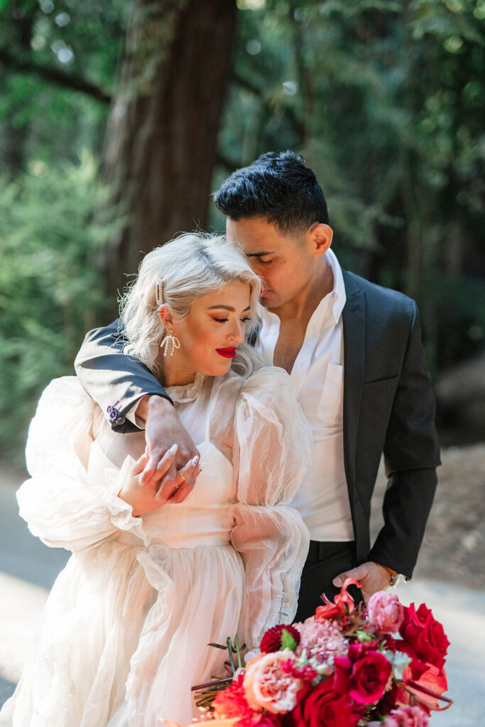 Editorial wedding portrait of bride and groom embracing in a California redwood forest with vibrant red bouquet photographed by a West Coast destination wedding photographer