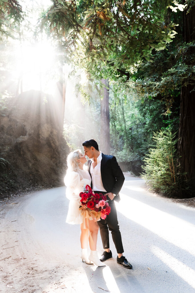 Couple sharing a kiss on a sunlit path through a California redwood forest with dramatic light rays photographed by a Las Vegas destination wedding photographer available across the West Coast