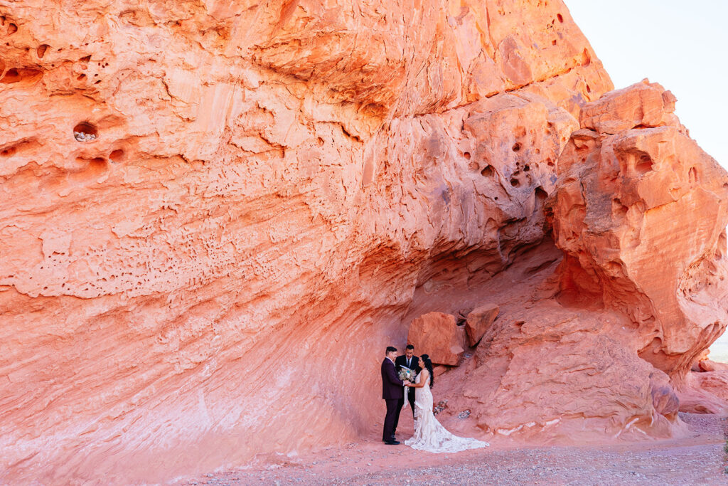Valley of Fire elopement photographer captures couple exchanging vows with their officiant beneath a towering red sandstone rock formation, bride in a fitted lace gown with a long train, groom in a burgundy suit, Nevada desert landscape at golden hour