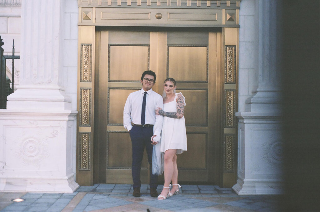 Film photo of newlywed couple posing in front of ornate gold courthouse doors, bride in short white dress with tattoos and groom in dress shirt and tie for intimate city elopement.
