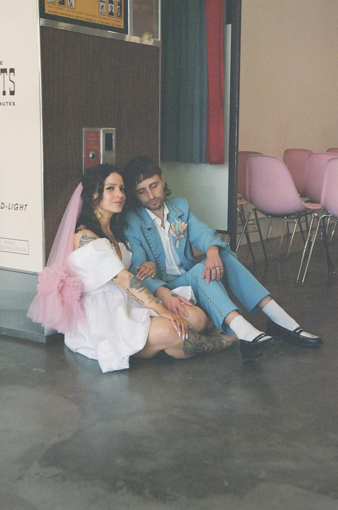Elopement photographer capturing a stylish couple seated on the floor of a vintage chapel, bride wearing a short white dress with pink veil and groom in a light blue suit with boutonniere.