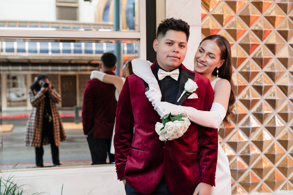 Las Vegas elopement photographer captures bride in white gloves holding roses wrapping her arms around groom in a burgundy velvet tuxedo, smiling outdoors in downtown Las Vegas — True Colors Creative